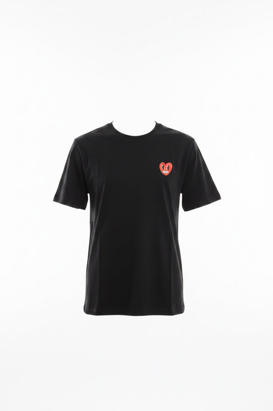 A black short-sleeve t-shirt with a red and white heart logo on the left chest, held up on a blue hanger by a hand in a warehouse or retail setting.