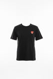 A black short-sleeve t-shirt with a red and white heart logo on the left chest, held up on a blue hanger by a hand in a warehouse or retail setting.